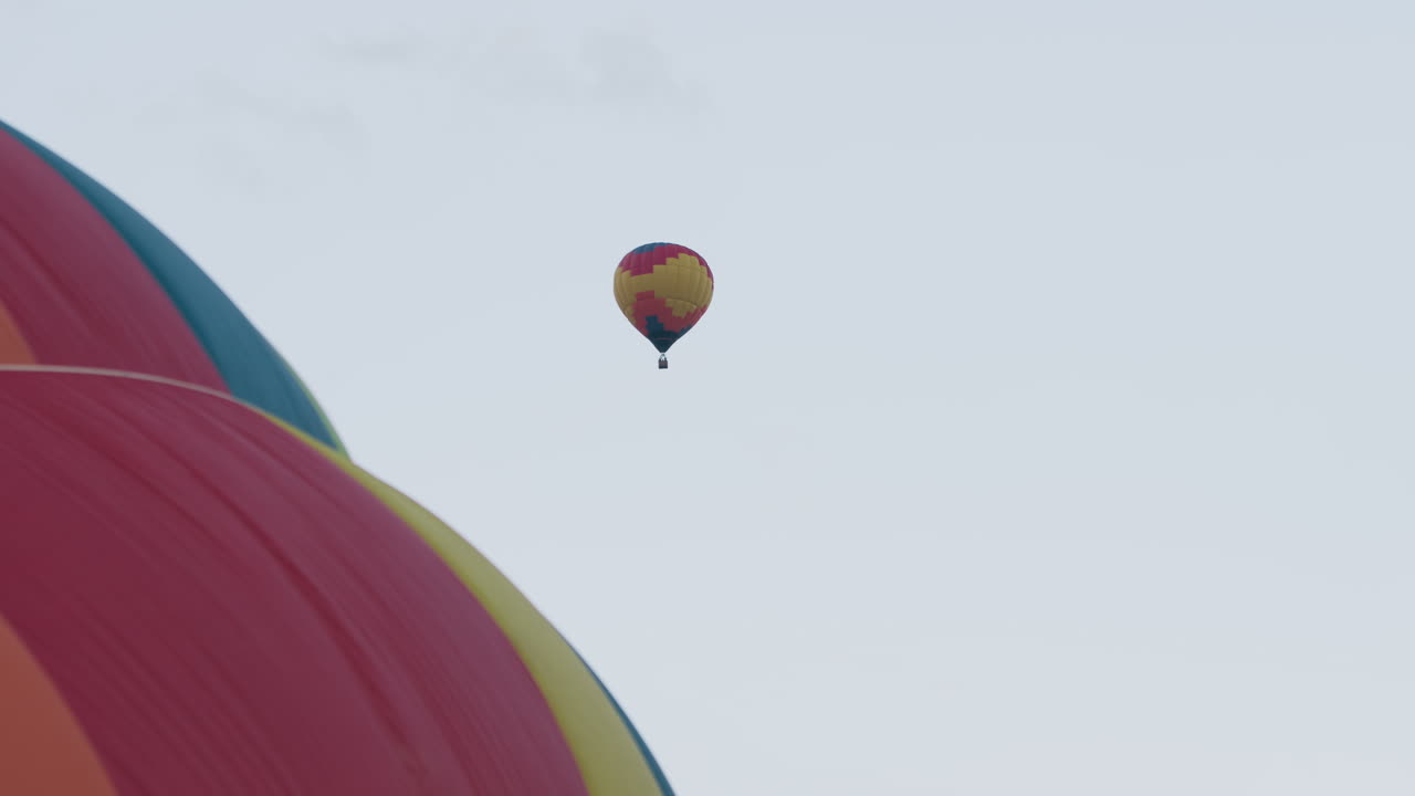 Close up hot air balloon envelope panels and basket gear in foreground with second balloon floating high in clear pastel sky, vivid red yellow and blue panels dominating left frame, serene airy scene
