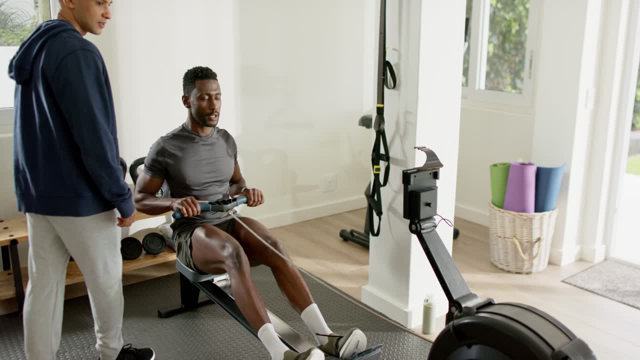 Coach cueing mid adult African American man wearing sportswear rowing on rower at gym for form