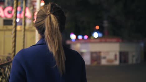 Back-view-of-young-attractive-woman-with-ponytail-walking-in-amusement-park-at-night,-turning-around-and-smiling-to-the-camera