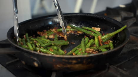 chef uses tongs to toss and stir sauteed green beans in pan, slow motion close up 4k