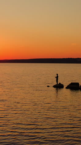 people fishing at sunset. beautiful orange sun on the evening sky. silhouette fisherman with fishing rod catching fish on the stone in water. men relax near the water. vertical video