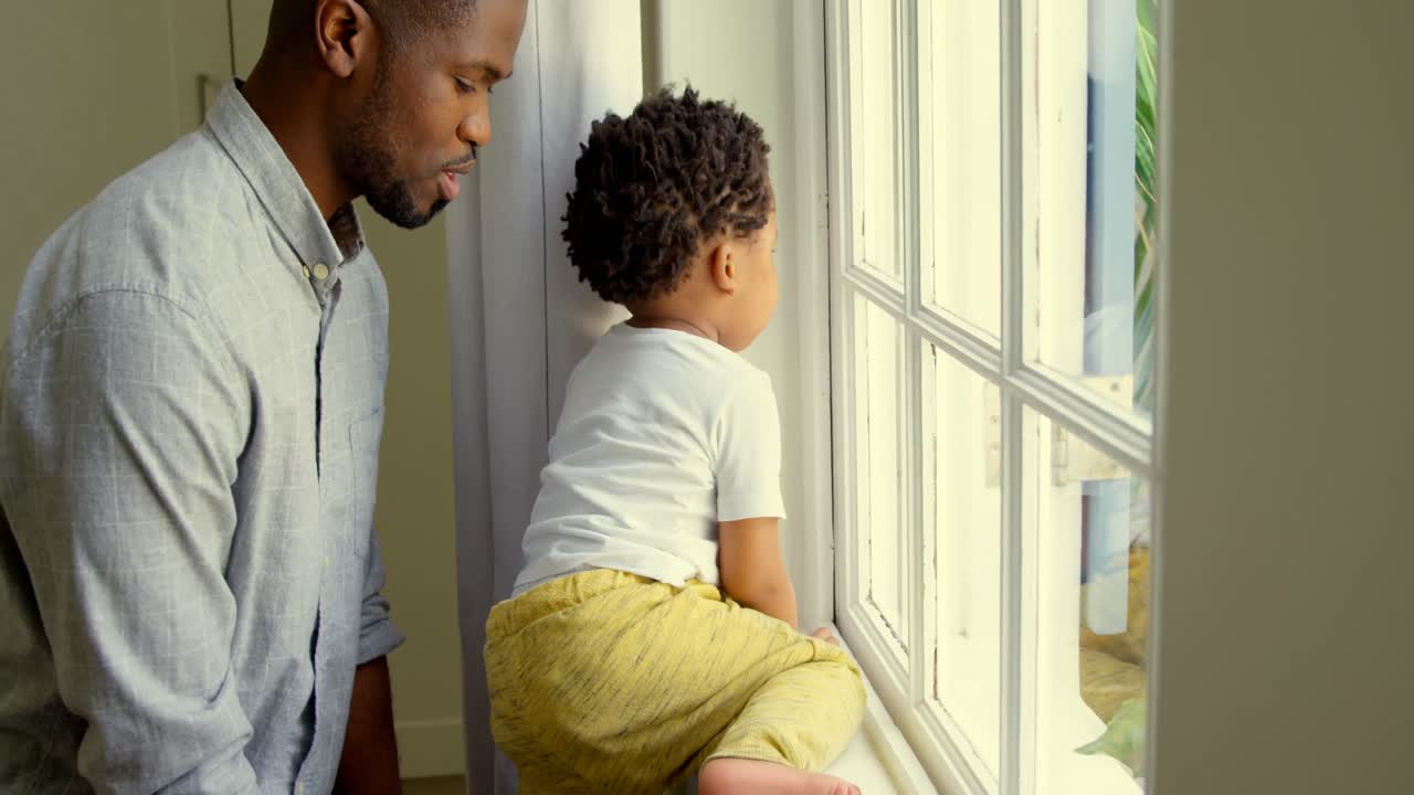 vista lateral de un joven padre negro jugando con su hijo en el alféizar de la ventana en una casa cómoda 4k