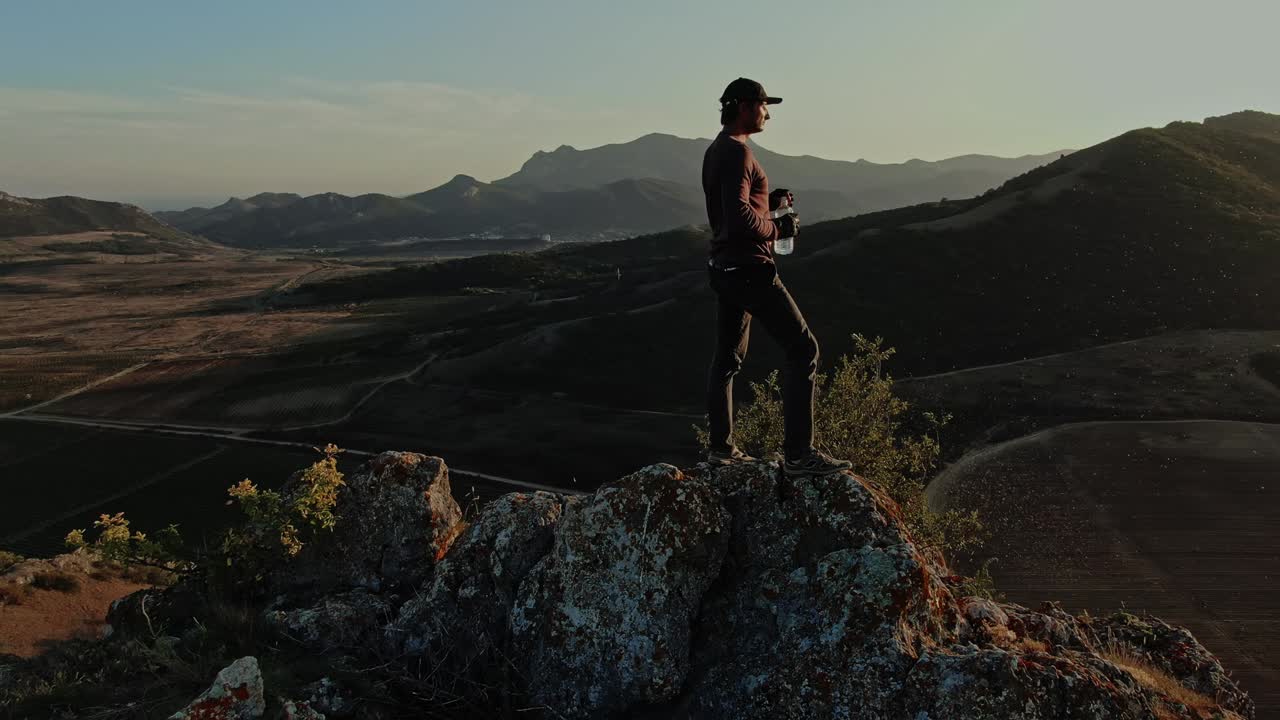 야구 모자를 입은 한 젊은이가 산의 꼭대기에 도달하여 끝없는 파란 하늘과 해가 지는 산의 풍경을 배경으로 투명한 플라스틱 병에서 마시고 있습니다.