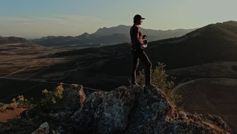 un joven con una gorra de béisbol ha llegado a la cima de la montaña y bebe de una botella de plástico transparente en el fondo de un paisaje montañoso del cielo azul sin fin y la puesta de sol. vista aérea. topografía por avión no tripulado con vuelo en círculo.