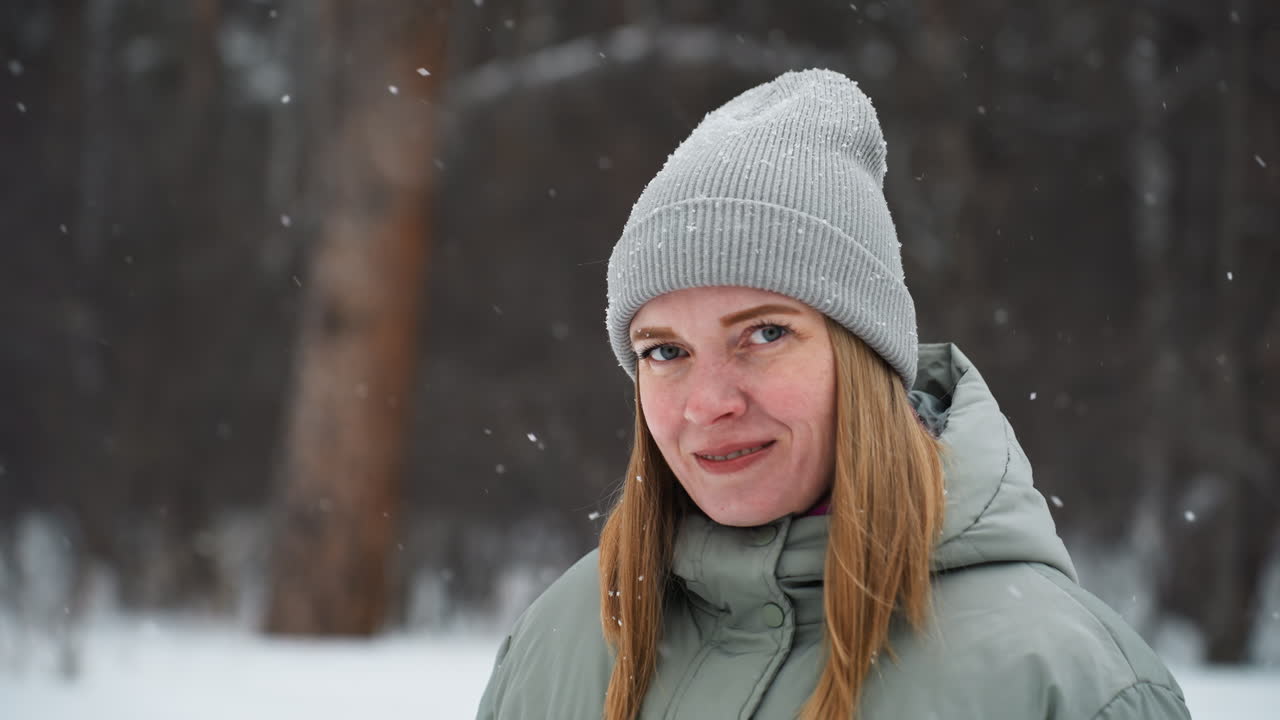Closeup of woman in gray beanie and winter jacket smiling gently while standing outdoors during snowfall, surrounded by blurred snowy forest background creating a peaceful and cozy winter