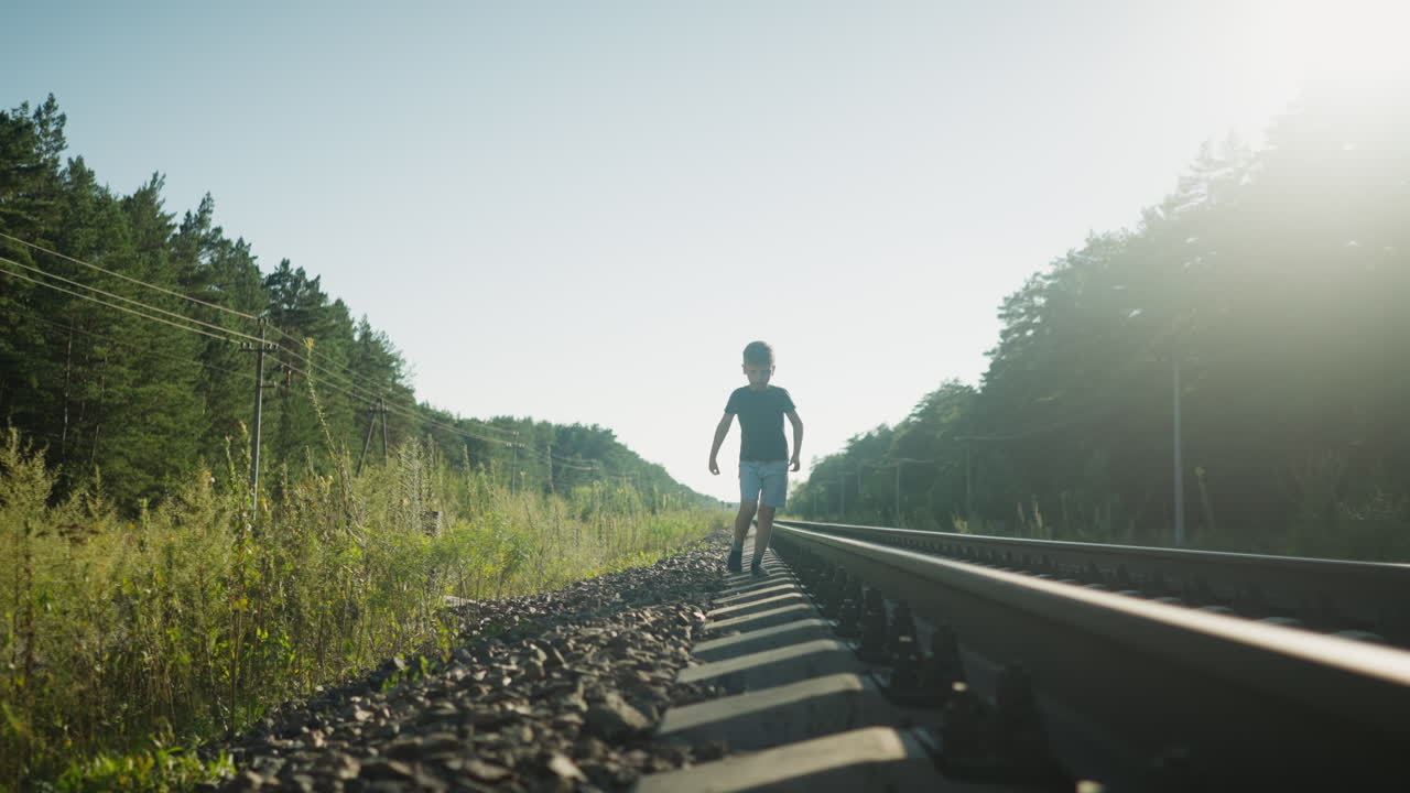 young boy walking rail track with careful steps across gravel ballast under glowing sunlight, framed by forest trees and power lines in quiet countryside