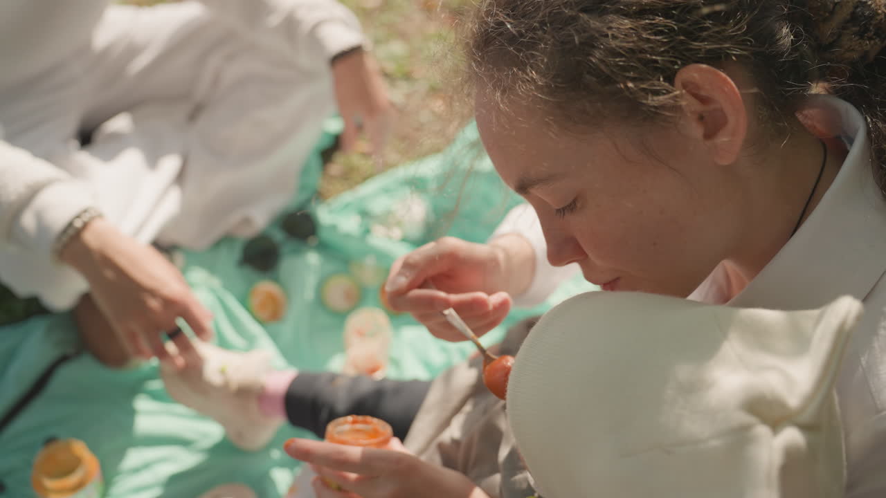 Figlia che assaggia la marmellata durante un picnic in famiglia, coperta color menta stesa sull'erba, mani dei genitori che offrono cucchiaio e vasetto, calda luce solare dorata, primo piano spontaneo dell'atto di nutrire e condividere, delicata cura, momento lento