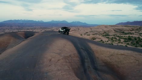 A-4K-tracking-drone-shot-of-a-lifted-jeep-off-roading-through-the-extreme-and-rocky-desert-landscape-near-Moab,-Utah,-with-the-snowy-rocky-mountains-towering-in-the-distance