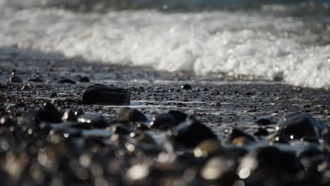 White-wash-froths-from-waves-crashing-on-pebble-beach,-glistening-in-slow-motion