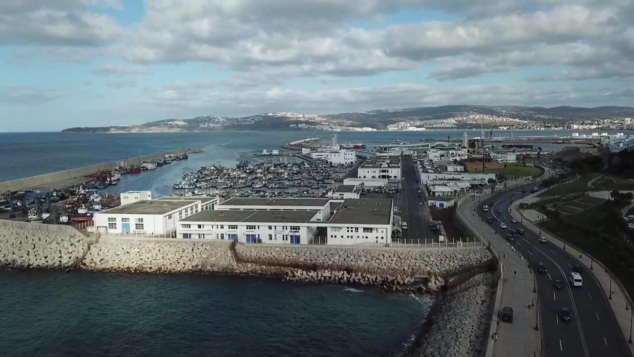 Letecký pohled dronem na pobřeží Tangeru, přístav a pobřežní silnice podél Středozemního moře pod dramatickými mraky. Filmová městská mořská krajina ideální pro cestovní a dokumentární projekty.
