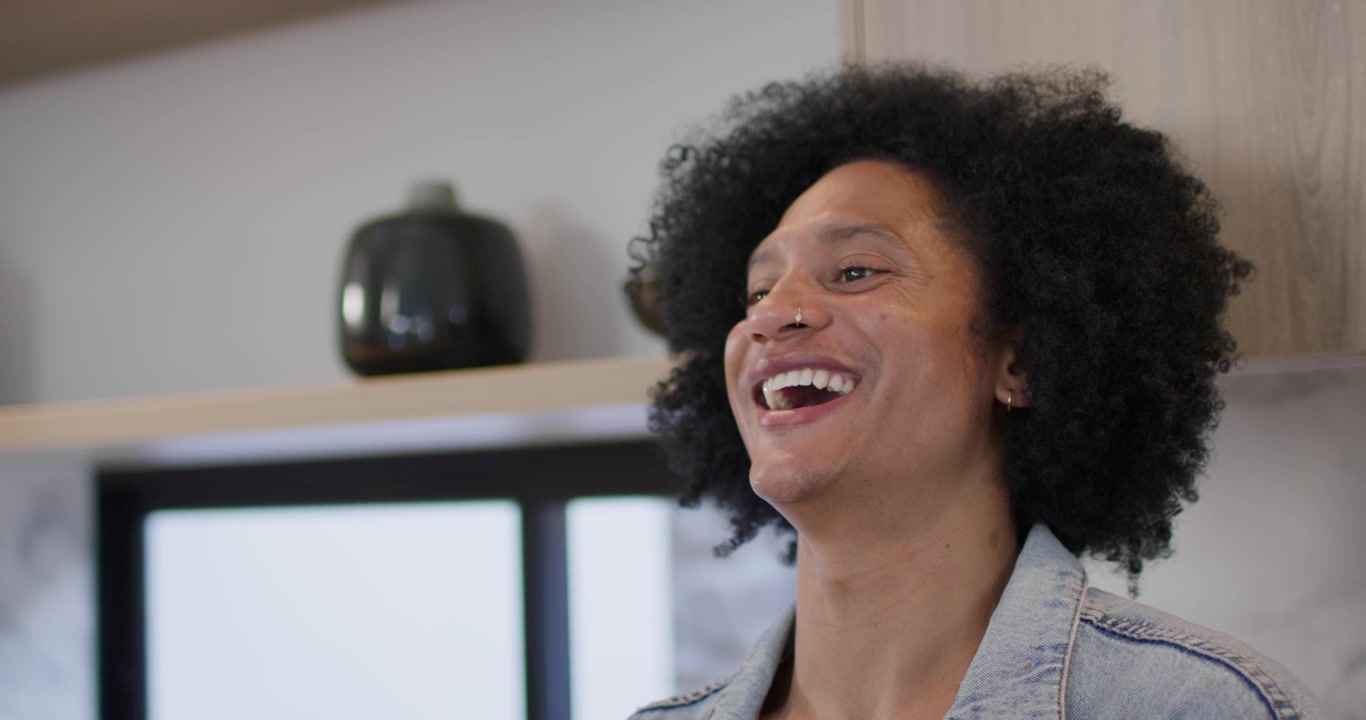Donna afroamericana che alza lo sguardo, sorridendo e ridendo, con una giacca di jeans e orecchini a cerchio, al bancone della cucina.