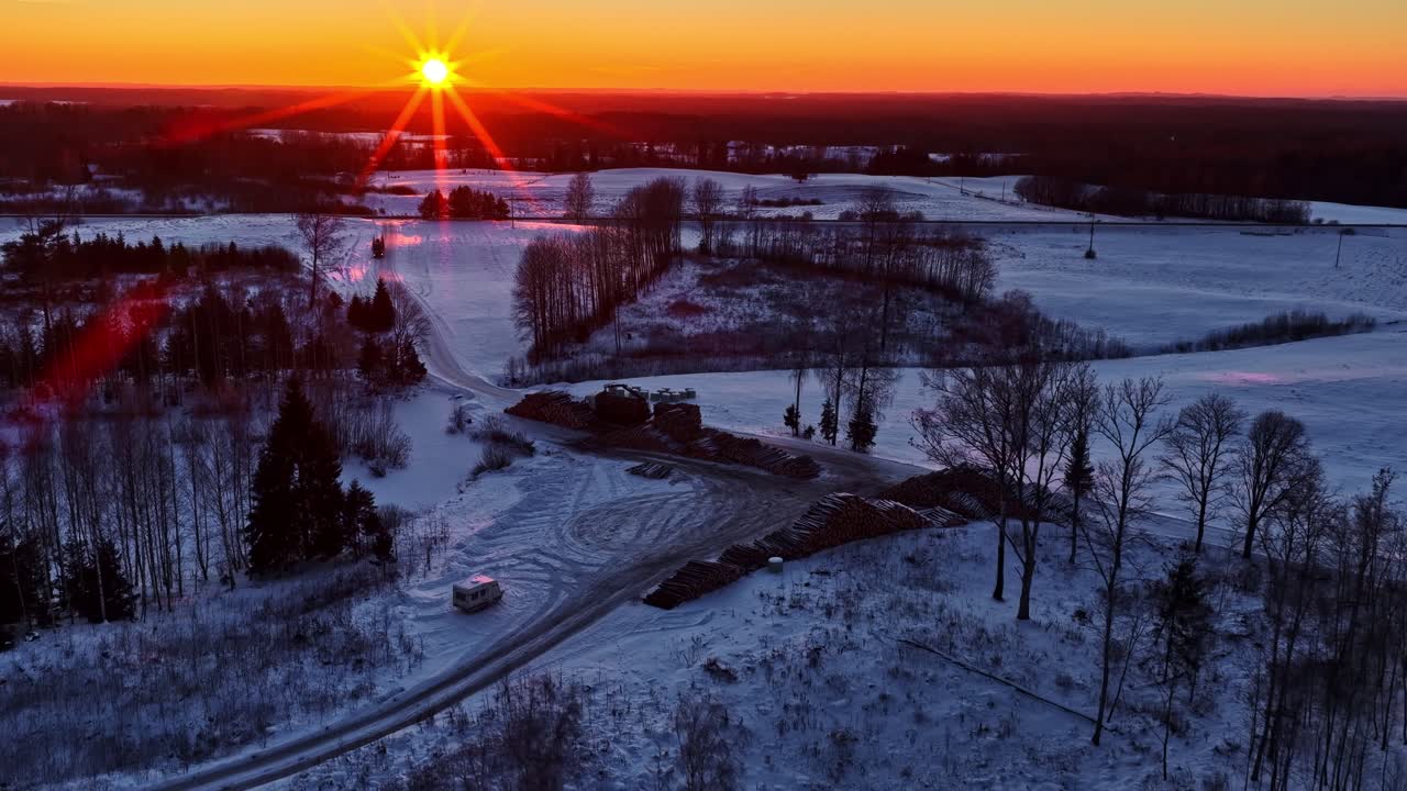 Toma de dron de ángulo alto de pilas organizadas de troncos de madera en un sitio de tala forestal durante un amanecer dorado de invierno en la campiña letona.