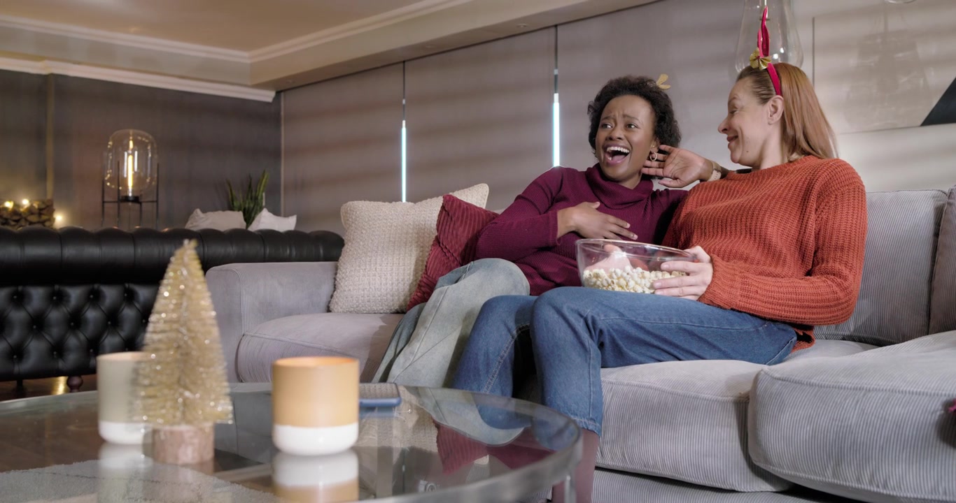 Mangfoldigt kvindepar i trøjer ser tv på sofaen, reagerer chokeret og sætter popcorn fra sig