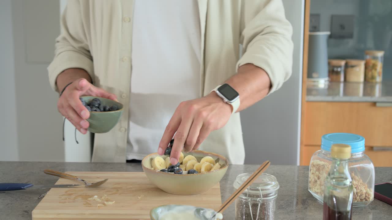 Mentre prepara la colazione, l'uomo aggiunge mirtilli da una ciotola sopra una banana, cospargendo semi sul bancone della cucina.