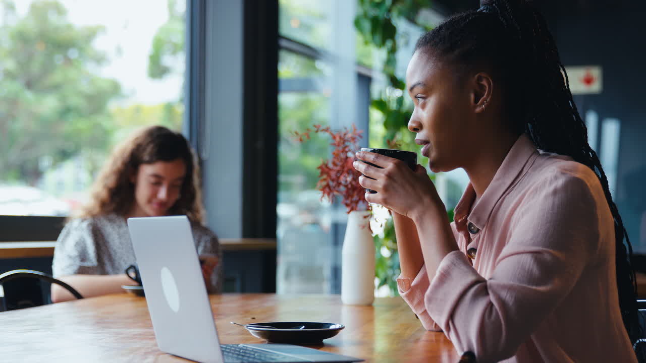 Jovem empresária com café trabalhando no notebook sentada em uma cafeteria com um colega ao fundo.