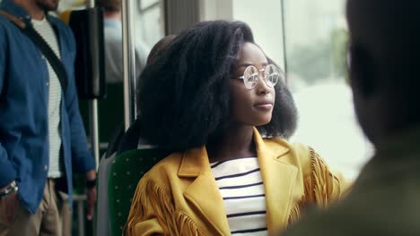 close up of the good looking and stylish young girl in glasses and with curly hair looking in the window while sitting in the tram, then turning face to the camera and smiling cheerfully