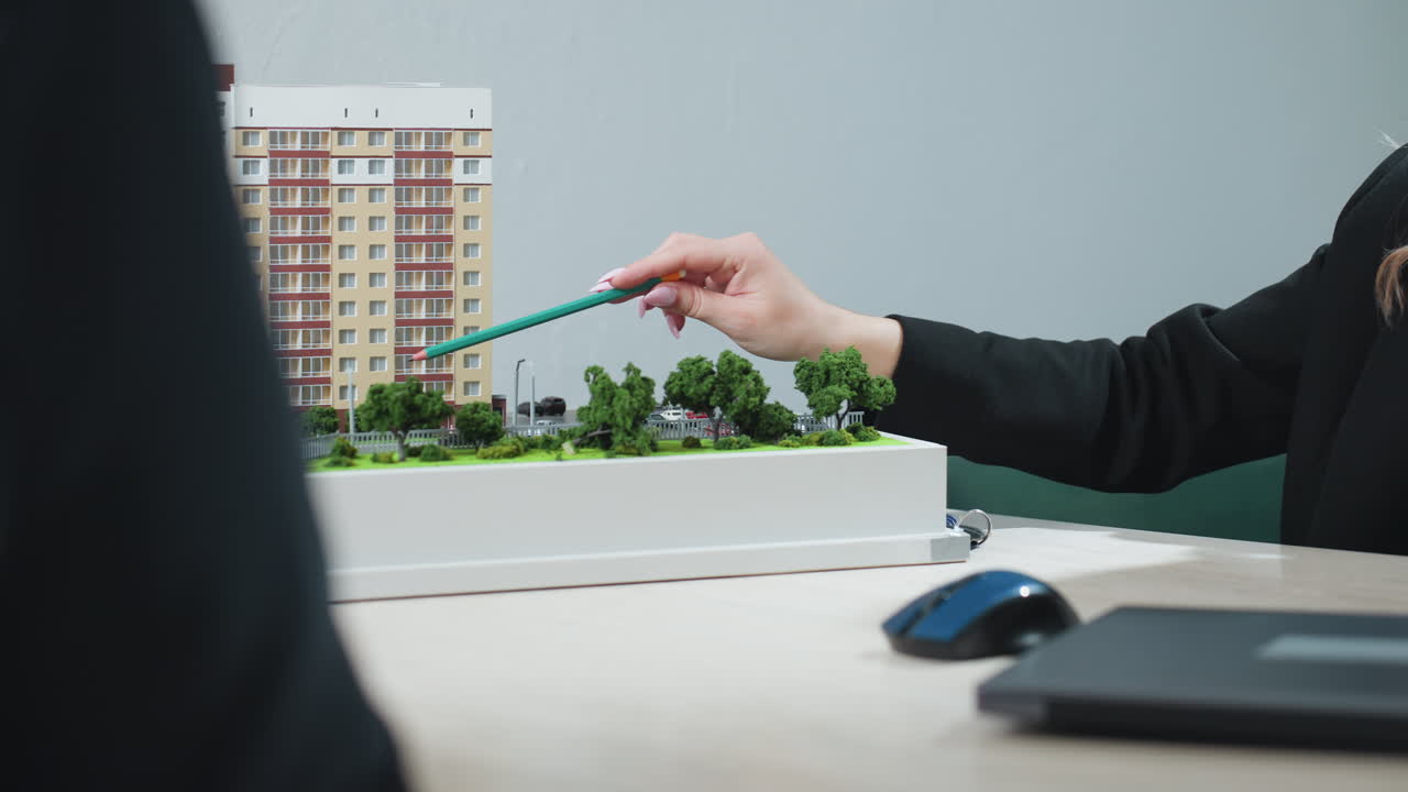 Real estate agent using pencil to explain features of residential building model to client during office consultation, with hands resting on desk, green trees surrounding miniature layout
