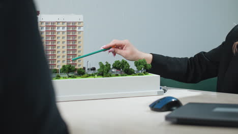real estate agent using pencil to explain features of residential building model to client during office consultation, with hands resting on desk, green trees surrounding miniature layout