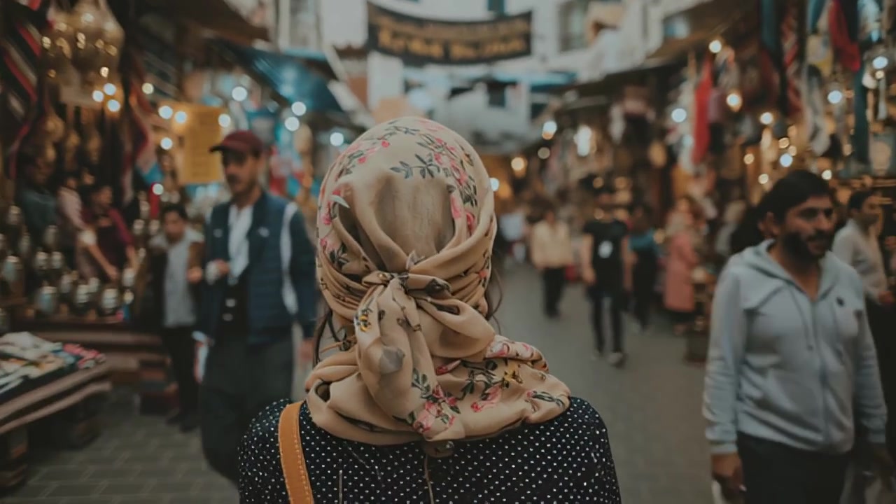 Cinematic rear view of a woman wearing a floral headscarf walking through a crowded bazaar street with shops, warm lights, and pedestrians in soft focus