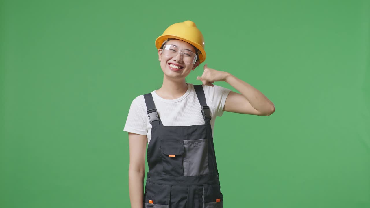 안경과 안전 헬을 입은 아시아 여성 노동자가 미소 짓고 녹색 스크린 배경 스튜디오에 서있는 동안 카메라에 제스처를 호출합니다.