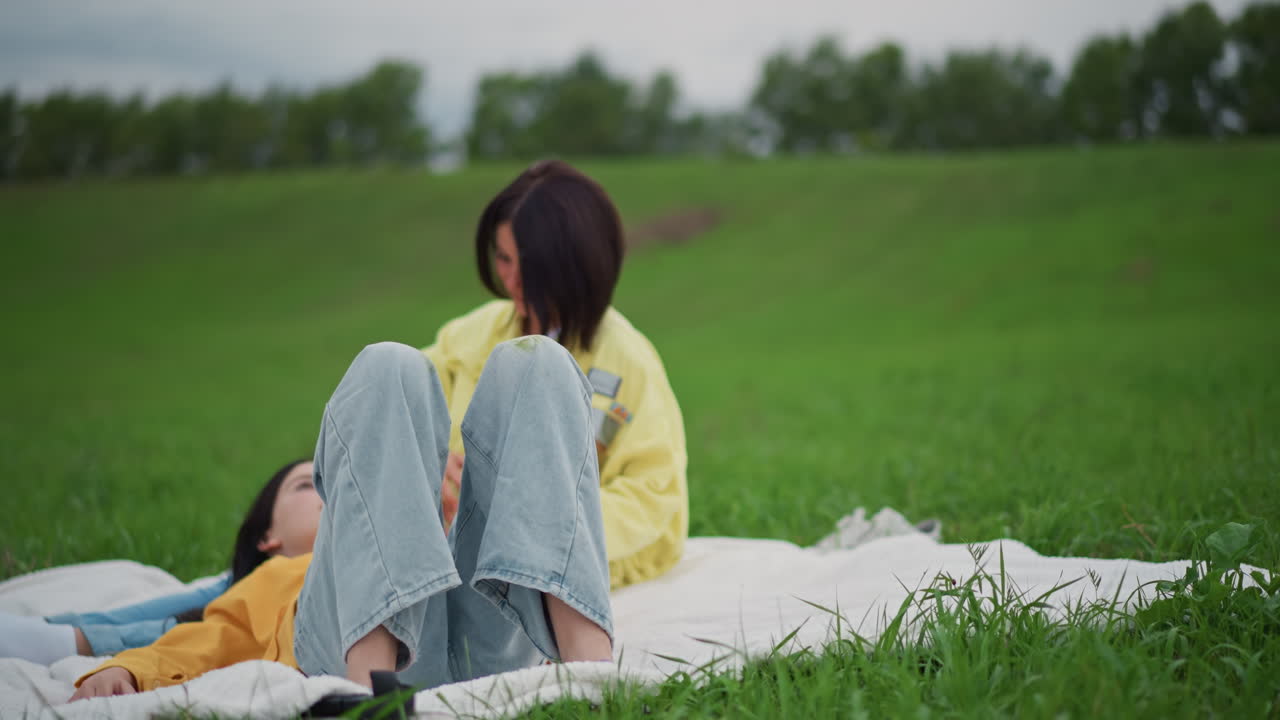 두 여성이 초록색 초원에서 담요를 덮고 휴식을 취하고 있어요. 한 명은 옆에 누워 있는 친구의 머리카락과 이마를 부드럽게 만져주고 있고, 은은한 빛이 감도는 가운데 캐주얼한 데님과 노란색 재킷을 입고 서로를 지지하며 친밀한 분위기를 연출하고 있어요.