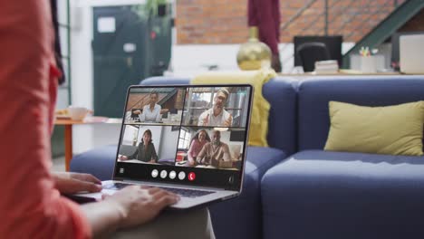 mujer biracial usando una computadora portátil para una llamada de video, con diversos colegas de negocios en la pantalla