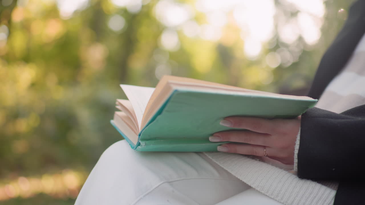 Lower view of elegant girl with legs crossed flipping pages of novel on lap, hands poised to reach drink beside bench, soft sunlight over park greenery, slow movement during quiet outdoor reading