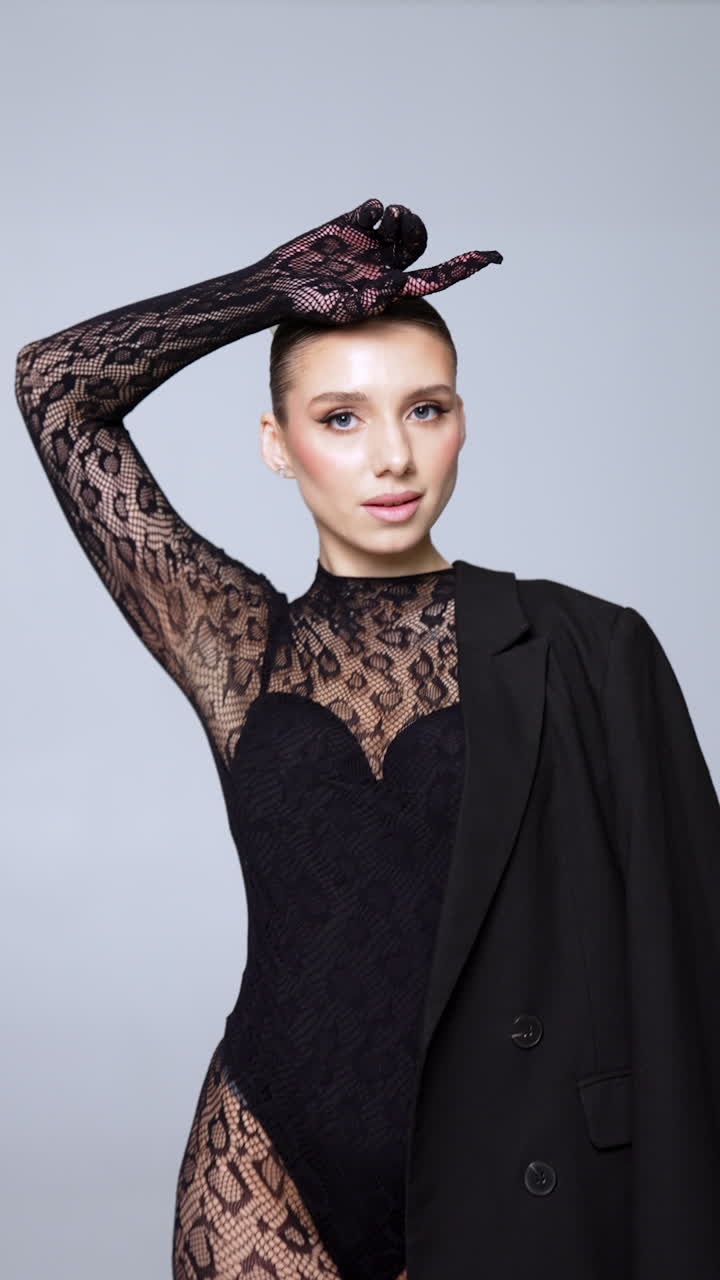 Beautiful model wearing black bodysuit and lace outfit poses in studio. Black jacket is hanging om female shoulder. Vertical video.
