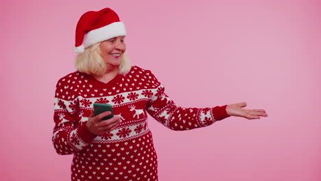 Grandmother-Santa-Christmas-sweater-with-mobile-phone-showing-pointing-empty-place,-advertising-area