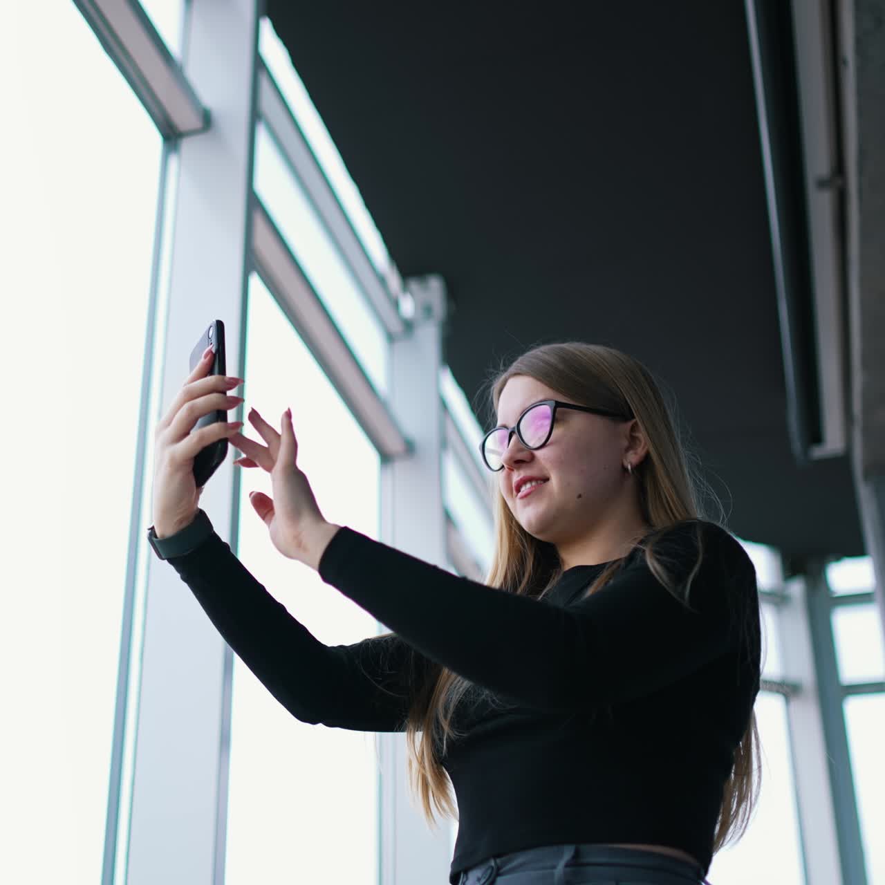 Beautiful woman doing photo of herself indoors. Attractive girl in glasses stands near the large windows and using mobile phone. View from below