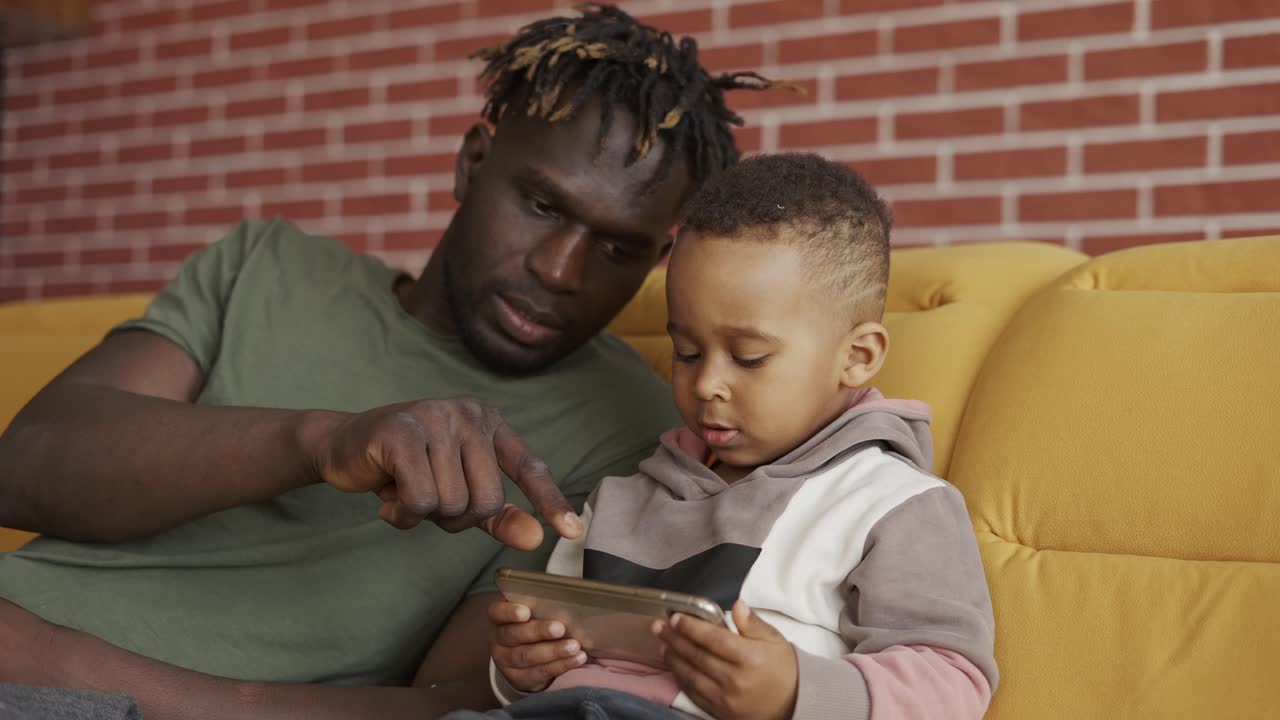 Ritratto di padre afroamericano seduto con il figlio sul divano - bambino che usa lo smartphone.