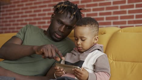 Portrait-of-african-american-father-sitting-with-son-on-sofa---kid-using-smartphone