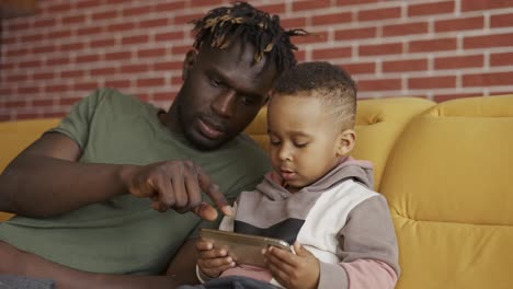 portrait of african american father sitting with son on sofa - kid using smartphone