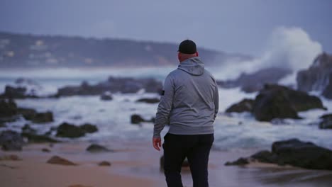 Male-walking-along-the-beach-while-giant-waves-crash-against-rocks