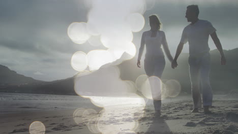 video compuesto de puntos de luz contra una pareja caucásica caminando por la playa