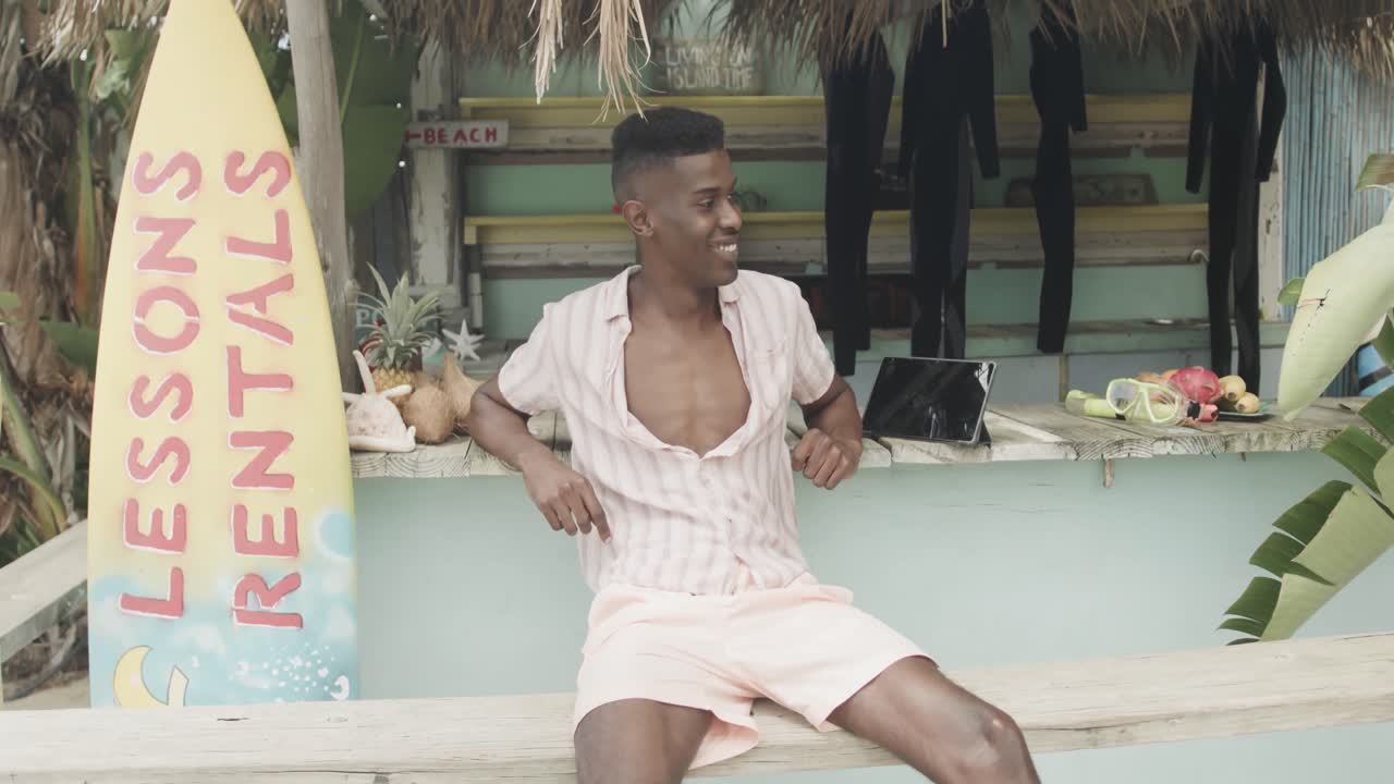 Glad afroamerikansk mand sidder og smiler udenfor en strandhytte med surfbrætsudlejning, slowmotion.
