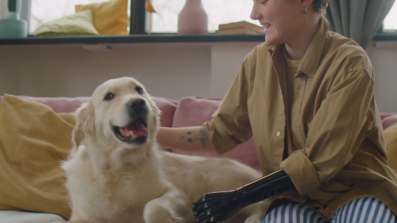 Young Woman with Prosthetic Arm Petting Playful Dog on Sofa