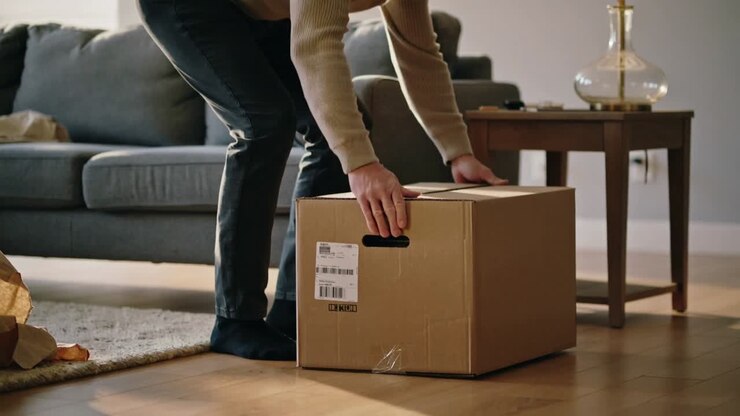 Carrying man in tan sweater entering living room shifting grip placing box orienting label outward