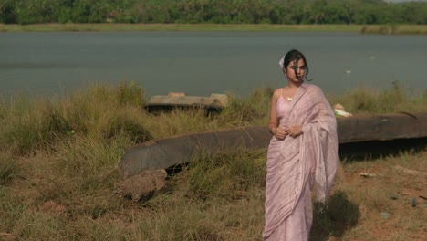 woman in pink saree standing by a river, looking over shoulder, with lush greenery in background