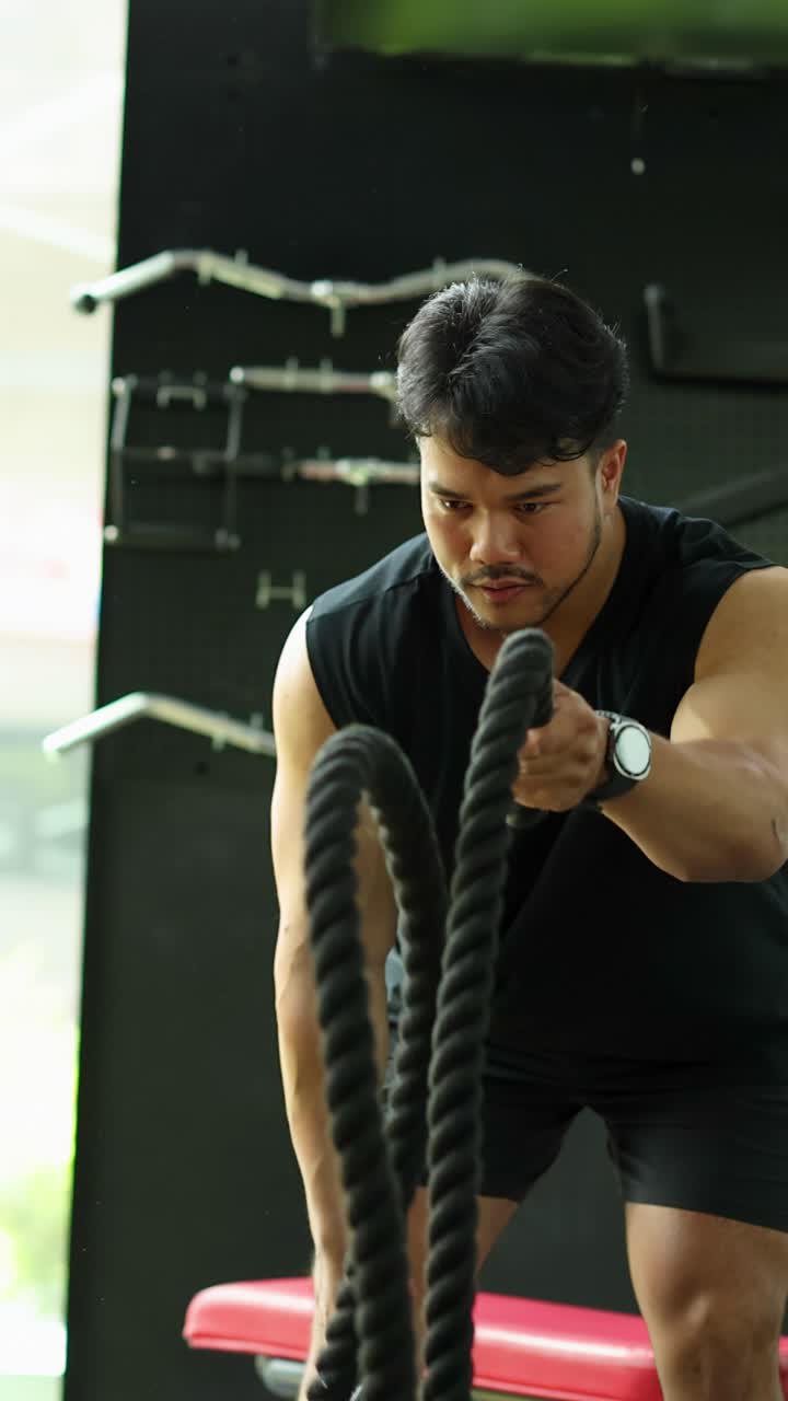 Homem atlético em roupa esportiva preta balança cordas navais em uma academia iluminada para treinamento funcional