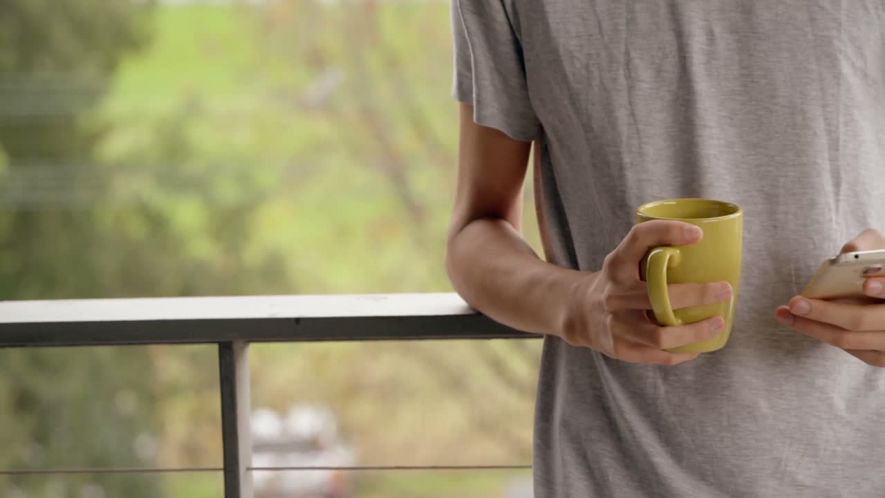 hombre usando teléfono móvil mientras sostiene una taza de café en el balcón 4k