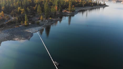 Aerial-shot-of-a-fishing-boat-near-dam-on-a-blue-alpine-lake-surrounded-by-pine-trees-and-mountains