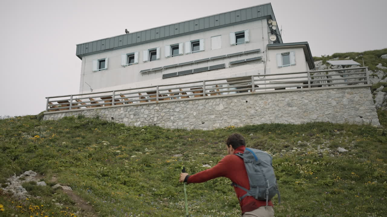 excursionista caminando por una colina hacia la cabaña de montaña en un día nublado de verano usando bastones de senderismo y vistiendo una chaqueta roja