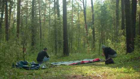 man squats holding flexible tent pole while woman stabilizes opposite end and aligns tent fabric beneath trees, sunlight softly glows through woods creating warm camping scene with gear