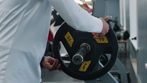 close up of person fixing weight plate onto barbell sleeve and securing with spring clip on rubber gym floor, showing hands positioning heavy chrome plate during workout preparation and setup