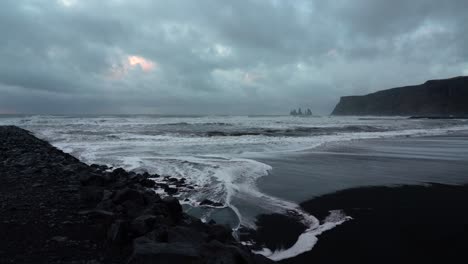 wide shot of black beach with reaching waves in vik,iceland during cloudy day - famous basalt sea stacks in background