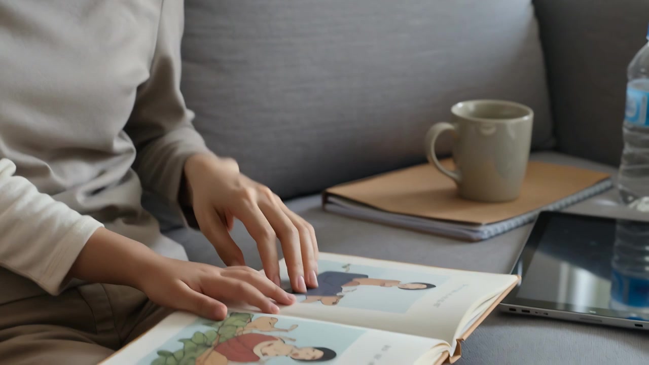 Prendendo un libro, madre e figlia asiatiche sfogliano le pagine, condividendo una storia su un divano grigio, con una tazza e un tablet.