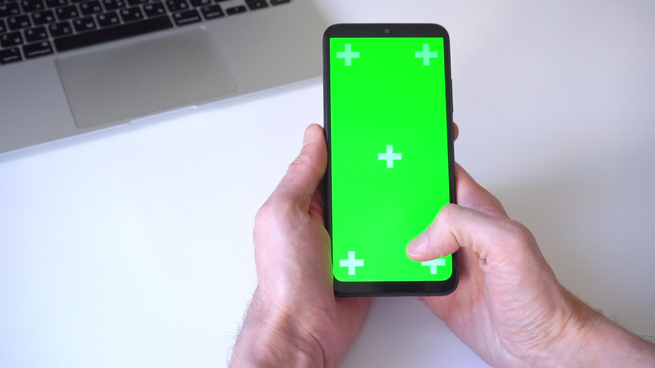 close-up. man holds smartphone both hands scrolls and presses his finger on green screen chromakey. on background of laptop and white table. concept of people, technology, social networks, web surfing