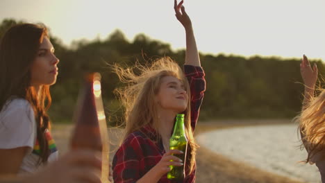 youg girls celebrate a birthday on the open air party with friends with beer and good mood. they dance in the summer evening near the river coast. this is carefree party at sunset.