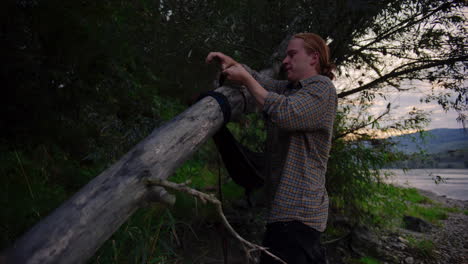 Handsome-young-man-with-hair-in-bun-wraps-hammock-straps-around-fallen-tree